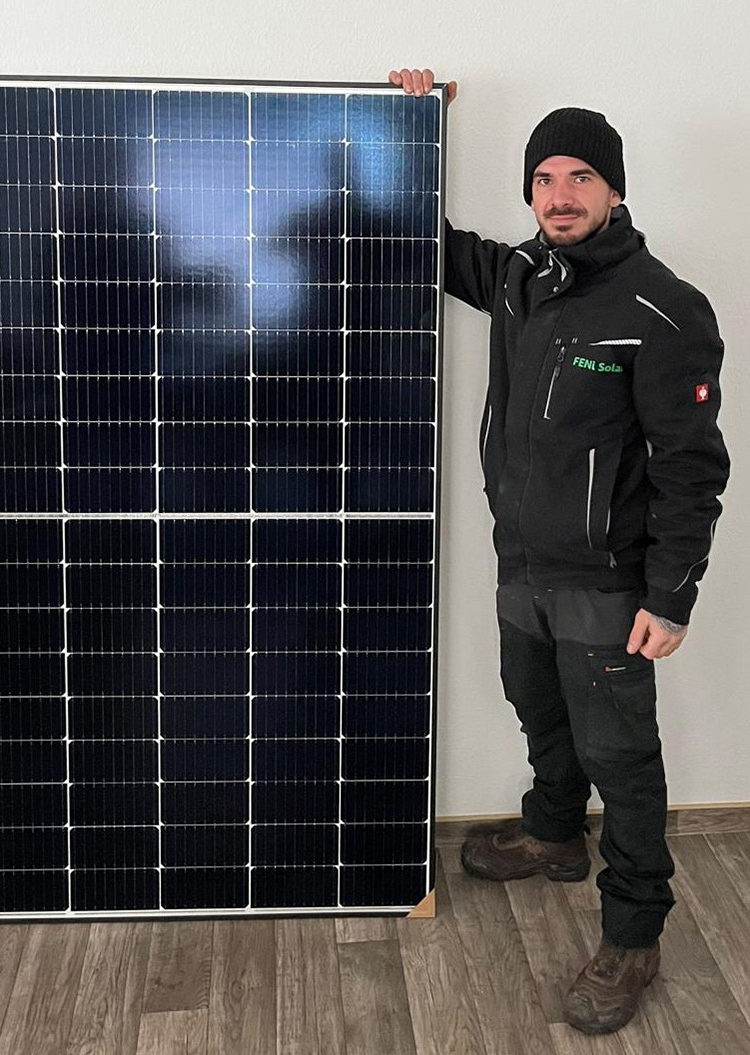 Ein lächelnder Mann in dunklem Arbeitsoutfit steht neben einem Solarpanel.