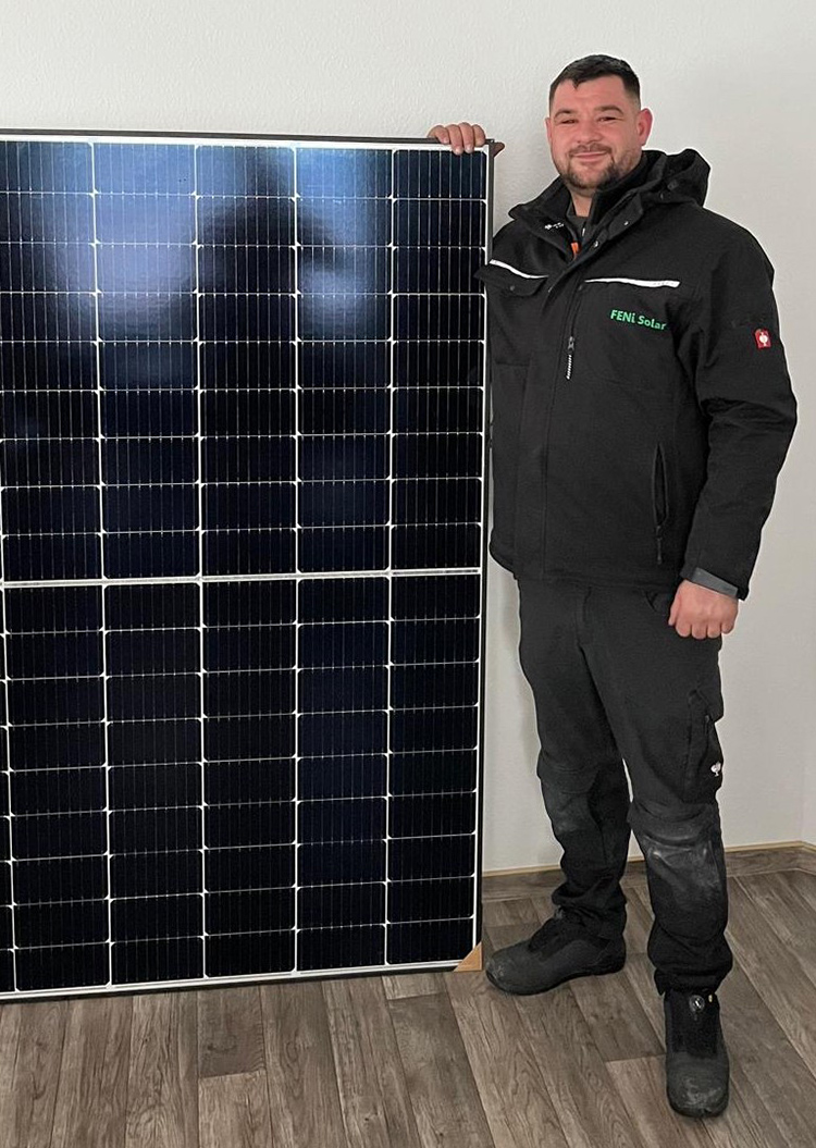 Ein Mann in schwarzer Kleidung lächelt neben einem großen Solarpanel.