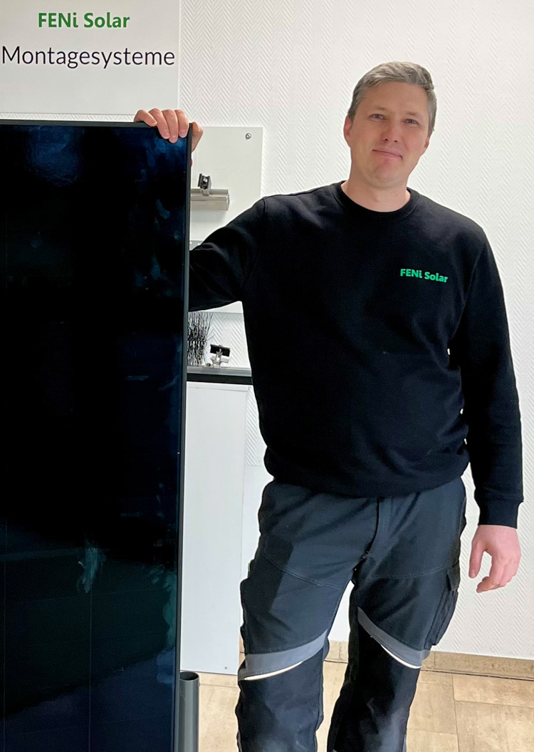 Ein Mann mit neutralem Gesichtsausdruck trägt dunkle Arbeitskleidung und steht neben einem Solarpanel