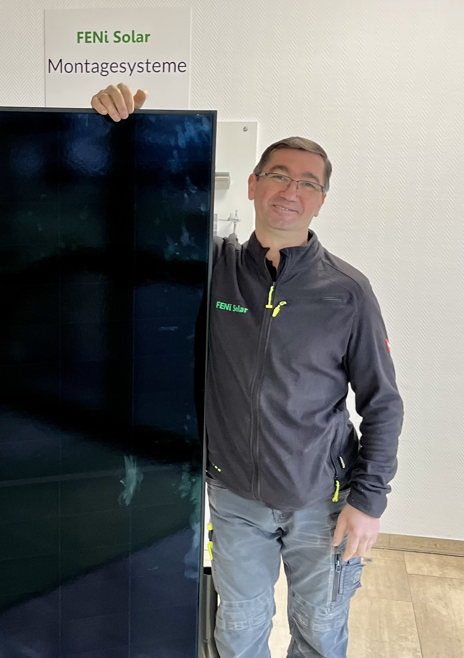 Ein lächelnder Mann mit Brille steht in grauer Jacke neben einem Solarmodul