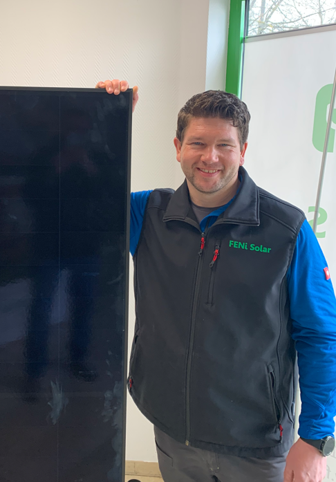 Ein lächelnder Mann mit dunklen Haaren trägt eine FENI Solar Weste und steht neben einem Solarpanel