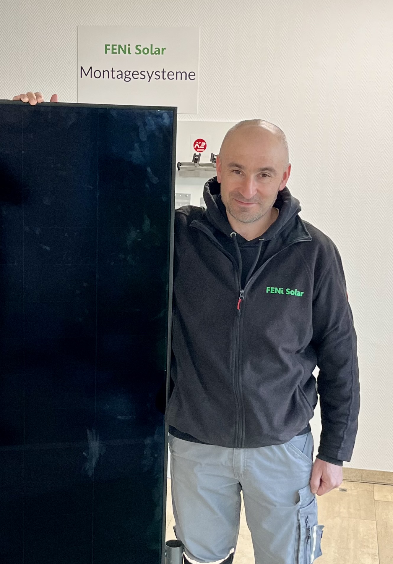 Ein Mann mit Glatze und grauer Jacke lächelt leicht und steht neben einem Solarpanel