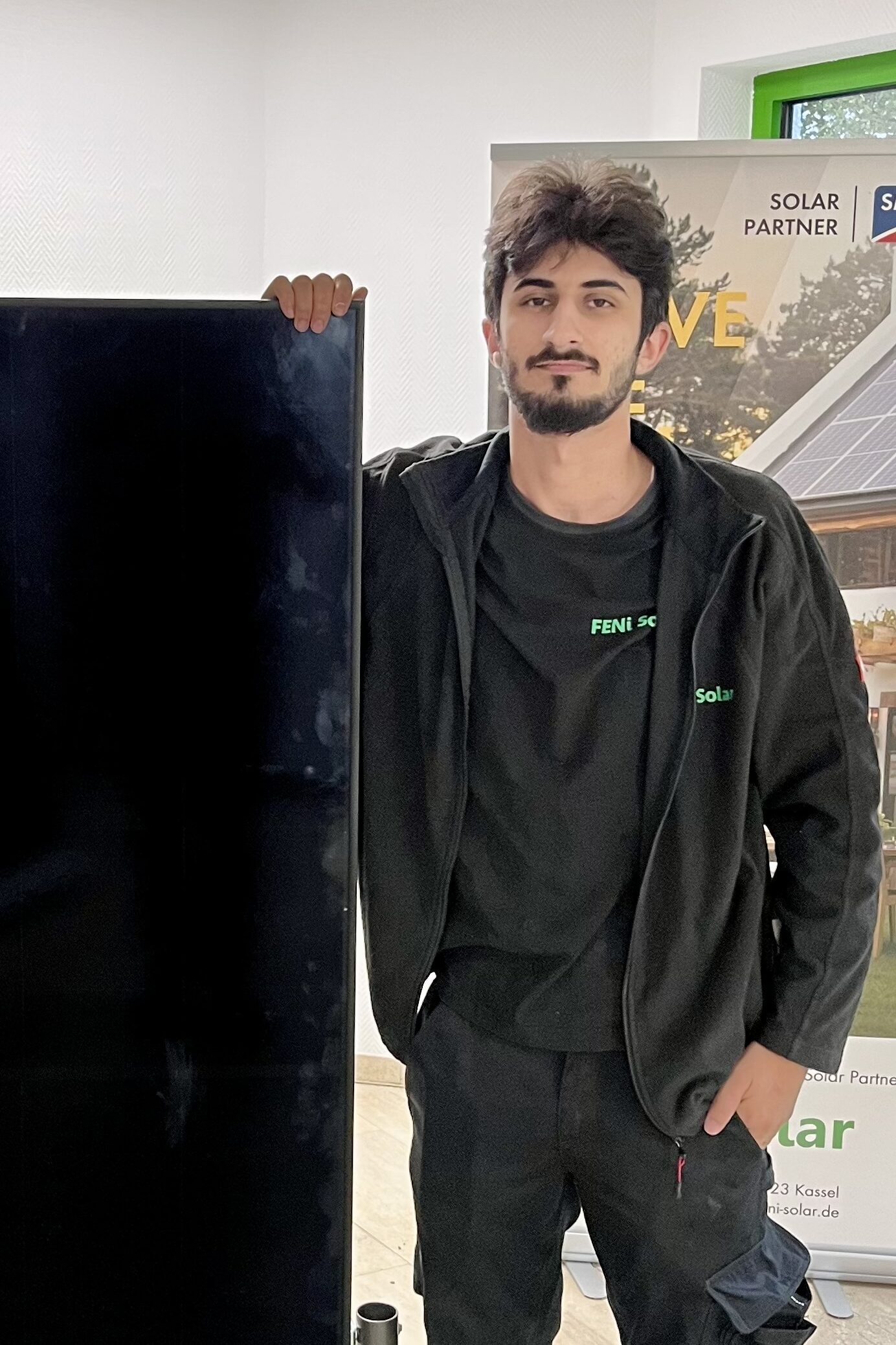 Junger Mann mit Bart in schwarzer Solar Arbeitskleidung haelt ein Solarmodul
