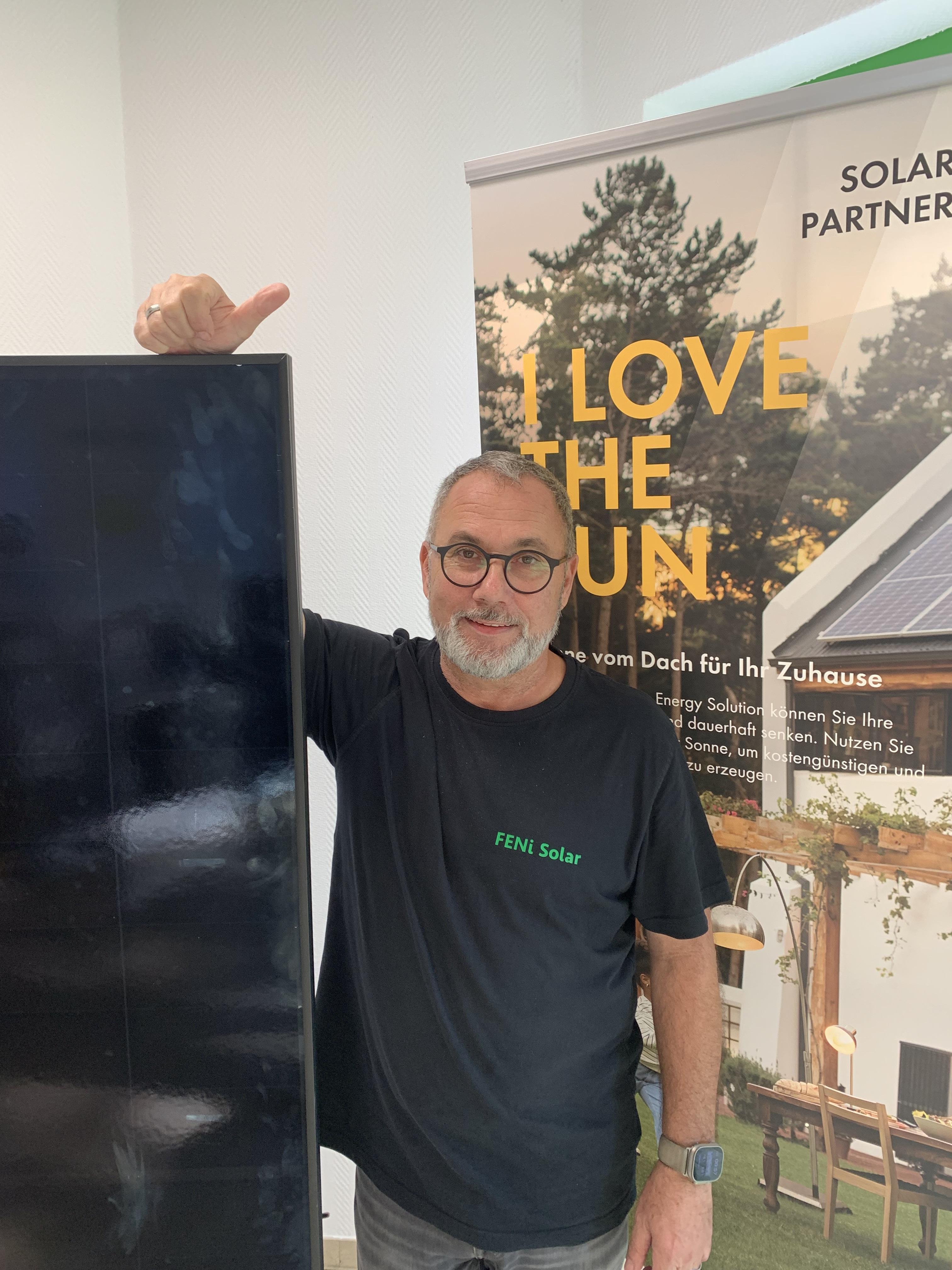 Laechelnder Mann mit Brille und Bart zeigt stolz auf ein Solarmodul